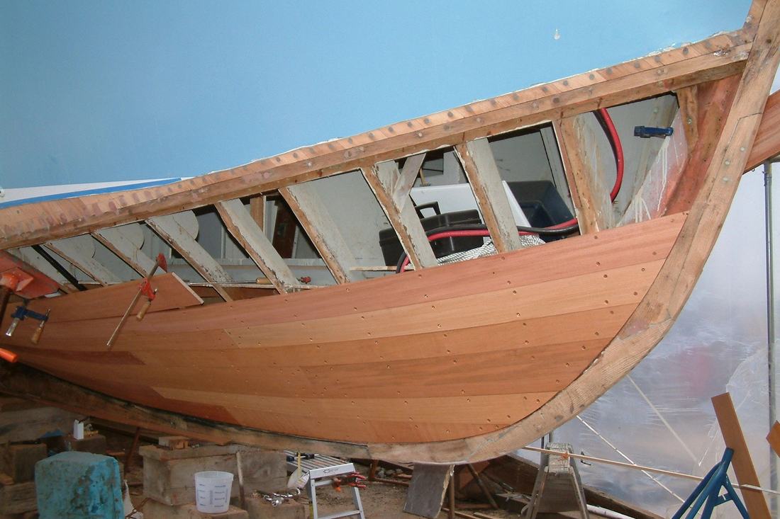 A wooden hull opened up on stands in the shed, frames and planking exposed during a refit.